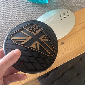 May include: A black and bronze motorcycle part with a Union Jack design and a diamond pattern. The part is held by a hand and sits on a wooden surface. A silver part with holes is behind it.