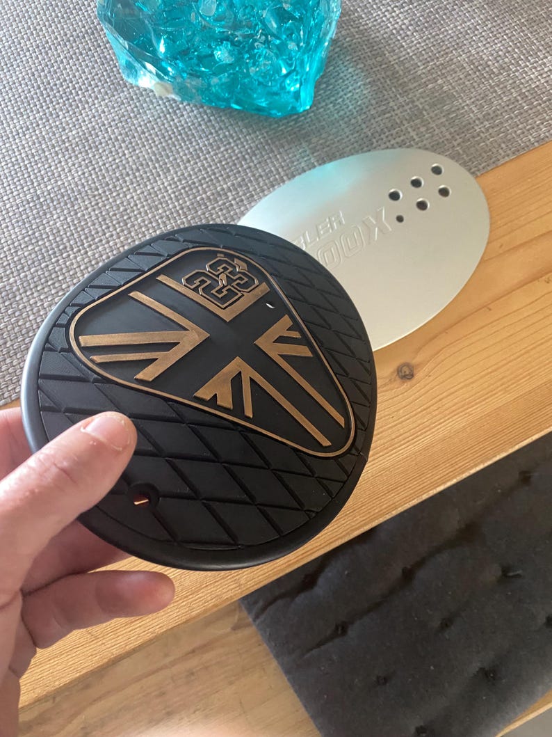 May include: A black and bronze motorcycle part with a Union Jack design and a diamond pattern. The part is held by a hand and sits on a wooden surface. A silver part with holes is behind it.