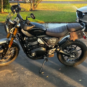 May include: Black and silver motorcycle with a brown seat and a Union Jack emblem. The motorcycle has a windshield, a small brown leather bag, and gold-colored front forks. The tires are black.