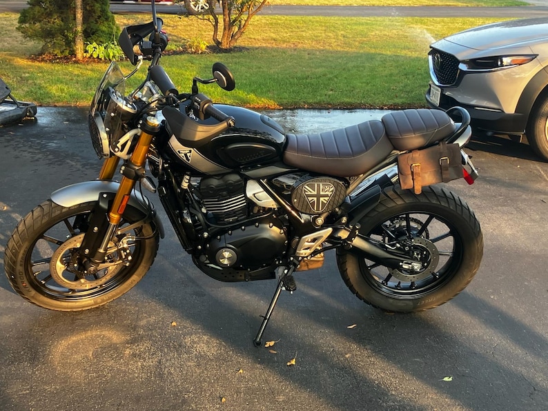 May include: Black and silver motorcycle with a brown seat and a Union Jack emblem. The motorcycle has a windshield, a small brown leather bag, and gold-colored front forks. The tires are black.