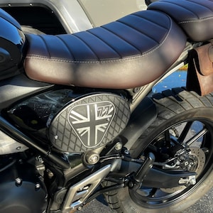 May include: A black motorcycle with a brown leather seat and a matching leather saddlebag. The side panel features a Union Jack design with the number 77. The motorcycle has a black tire and a silver metal frame.