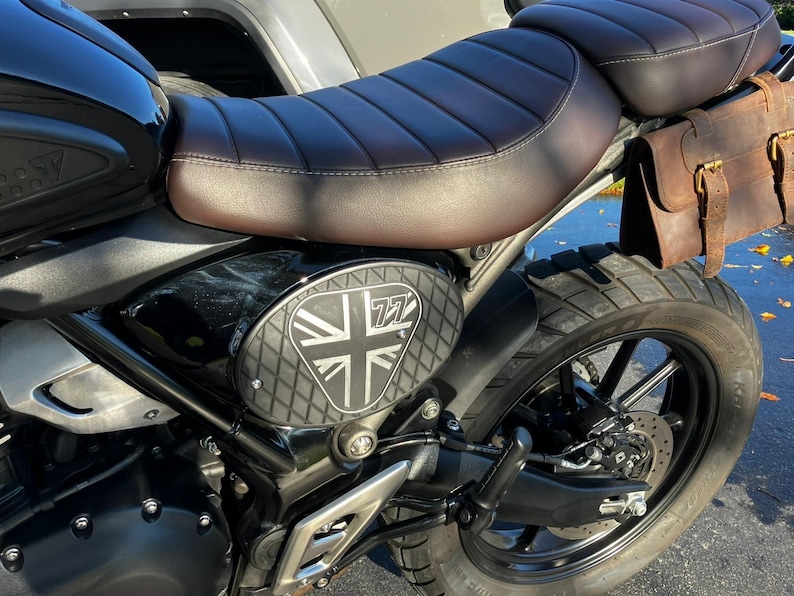 May include: A black motorcycle with a brown leather seat and a matching leather saddlebag. The side panel features a Union Jack design with the number 77. The motorcycle has a black tire and a silver metal frame.