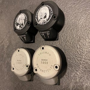 May include: Four circular motorcycle reservoir covers. Two black covers feature a silver anime-style character emblem and the text CYBERPUNK. Two silver covers are embossed with the text TRIUMPH SINCE 1902 MOTORCYCLES and have two screw holes.