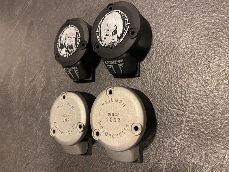 May include: Four circular motorcycle reservoir covers. Two black covers feature a silver anime-style character emblem and the text CYBERPUNK. Two silver covers are embossed with the text TRIUMPH SINCE 1902 MOTORCYCLES and have two screw holes.
