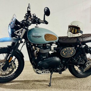 Może przedstawiać: Motocykl w stylu vintage z jasno niebieskim zbiornikiem paliwa i brązowym sk&oacute;rzanym siedzeniem. Motocykl ma czarne opony, biały kask z goglami i logo Triumph. Na zdjęciu widać r&oacute;wnież czerwoną gaśnicę.