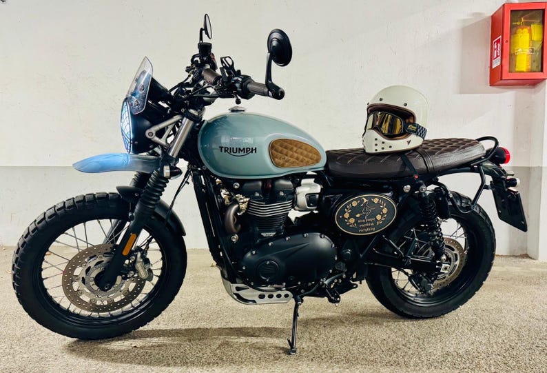 Może przedstawiać: Motocykl w stylu vintage z jasno niebieskim zbiornikiem paliwa i brązowym sk&oacute;rzanym siedzeniem. Motocykl ma czarne opony, biały kask z goglami i logo Triumph. Na zdjęciu widać r&oacute;wnież czerwoną gaśnicę.