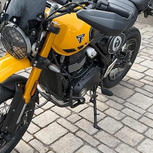 May include: A yellow Triumph motorcycle with black accents. The motorcycle has a black windscreen, headlight guard, and seat. The Triumph logo is visible on the fuel tank. The bike is parked on a brick surface.