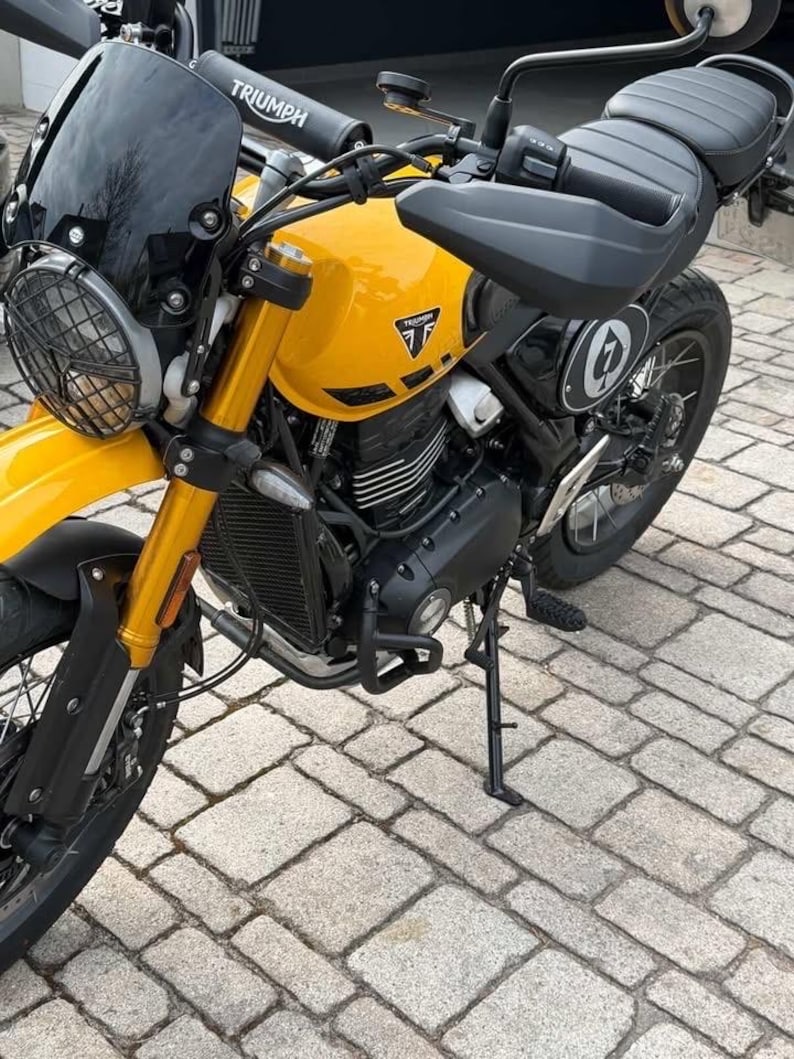May include: A yellow Triumph motorcycle with black accents. The motorcycle has a black windscreen, headlight guard, and seat. The Triumph logo is visible on the fuel tank. The bike is parked on a brick surface.