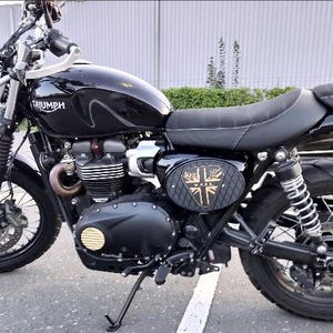 Może przedstawiać: Czarny motocykl Triumph z okrągłym reflektorem, czarnym siedzeniem i emblematem Union Jack z boku. Motocykl ma czarny zbiornik paliwa i silnik i jest zaparkowany na utwardzonej nawierzchni.