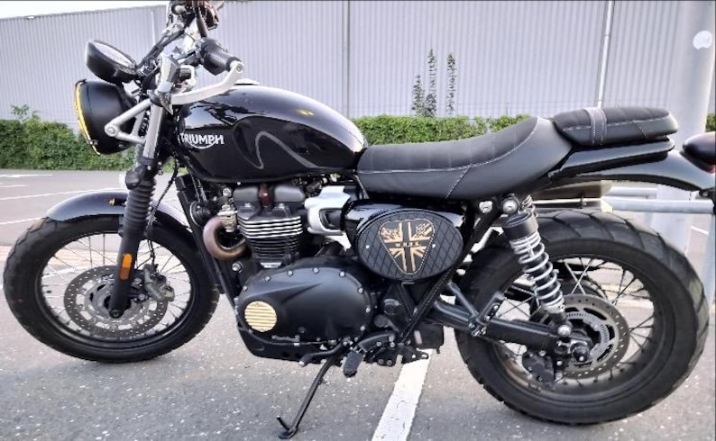 Może przedstawiać: Czarny motocykl Triumph z okrągłym reflektorem, czarnym siedzeniem i emblematem Union Jack z boku. Motocykl ma czarny zbiornik paliwa i silnik i jest zaparkowany na utwardzonej nawierzchni.