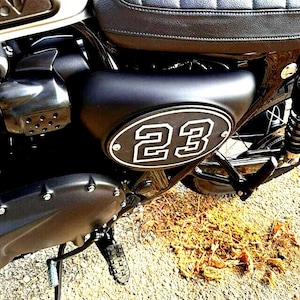 May include: A matte black motorcycle side panel featuring an oval-shaped racing number plate with the white number 23 outlined in black. The panel is positioned below a black quilted leather seat and above a black engine casing with visible bolts.