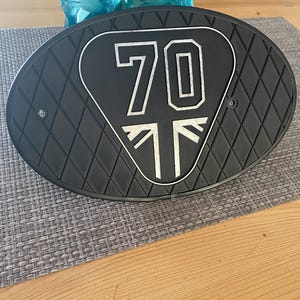 Może przedstawiać: Czarna owalna część motocykla z powierzchnią w romby. Środek przedstawia biały wz&oacute;r z numerem "70" i stylizowanym Union Jackiem. Część leży na szarej macie i drewnianym stole.