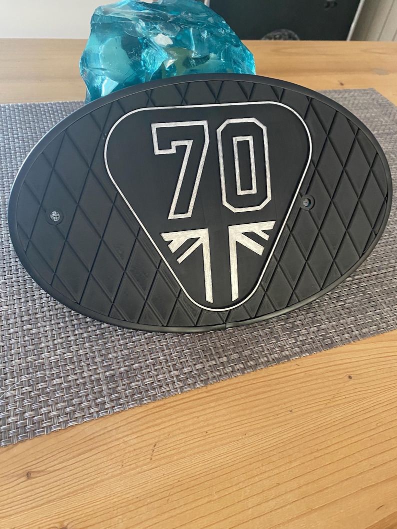 Może przedstawiać: Czarna owalna część motocykla z powierzchnią w romby. Środek przedstawia biały wz&oacute;r z numerem "70" i stylizowanym Union Jackiem. Część leży na szarej macie i drewnianym stole.