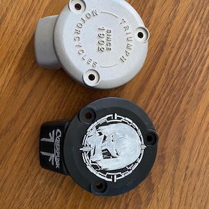 May include: Two circular metal engine covers with three mounting holes each. The top cover is silver with the embossed text TRIUMPH MOTORCYCLES SINCE 1902. The bottom cover is black with a white graphic of a helmet and the text CYBERPUNK.