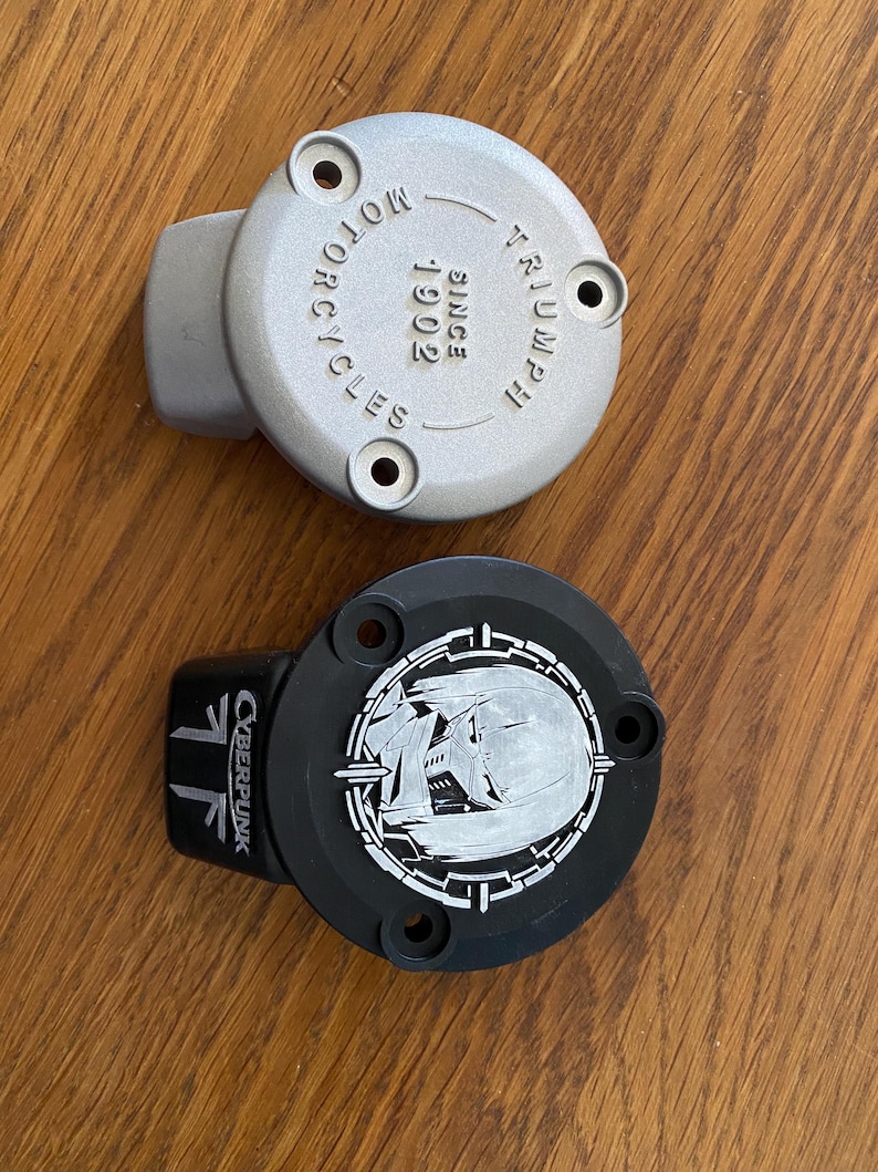 May include: Two circular metal engine covers with three mounting holes each. The top cover is silver with the embossed text TRIUMPH MOTORCYCLES SINCE 1902. The bottom cover is black with a white graphic of a helmet and the text CYBERPUNK.