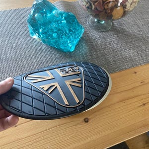 May include: A black and gold motorcycle part with a Union Jack design. The part has a textured surface and a gold emblem. A blue glass sculpture and a glass bowl with dried flowers are in the background.