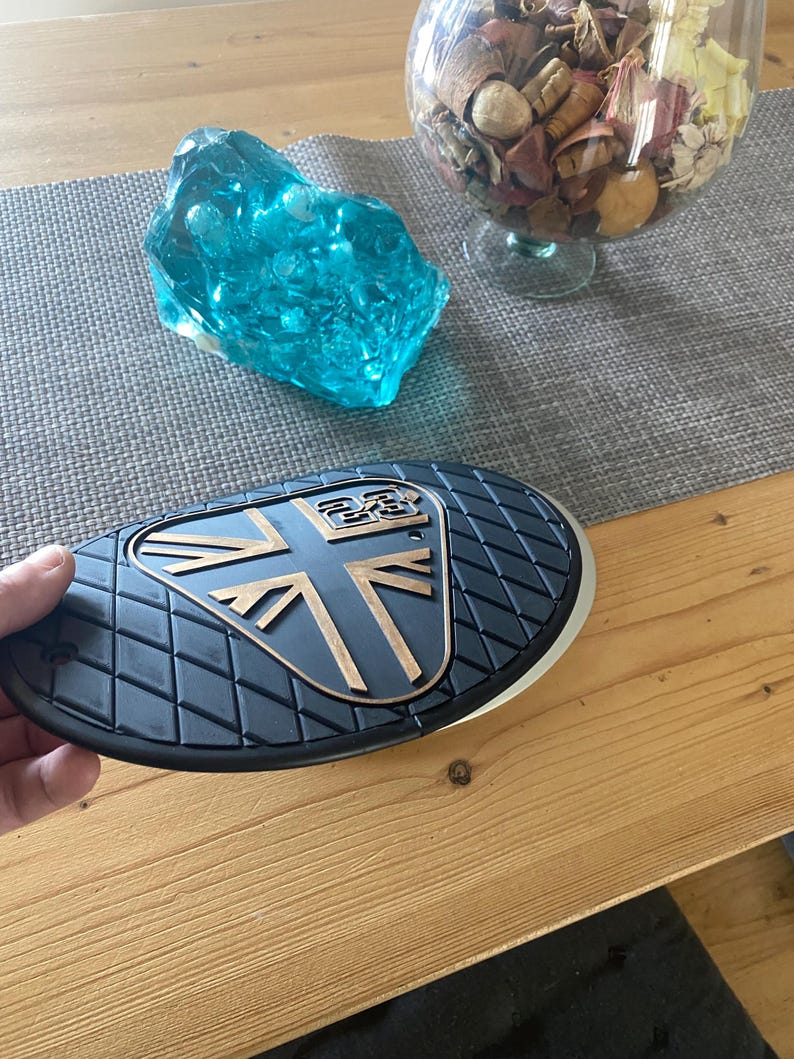 May include: A black and gold motorcycle part with a Union Jack design. The part has a textured surface and a gold emblem. A blue glass sculpture and a glass bowl with dried flowers are in the background.