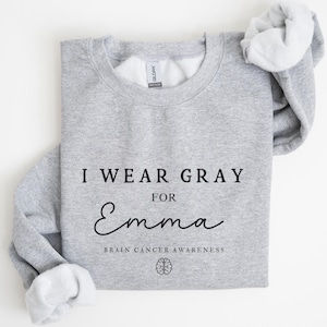 Puede incluir: Sudadera gris deportiva con el texto negro "I WEAR GRAY FOR Emma". La sudadera también incluye el texto "BRAIN CANCER AWARENESS" y un gráfico de cerebro. Las mangas están dobladas, revelando el interior blanco.