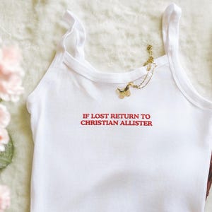 Könnte beinhalten: Ein weißes ärmelloses Tanktop mit rotem Sticktext, der IF LOST RETURN TO CHRISTIAN ALLISTER lautet. Das Kleidungsstück hat einen Rundhalsausschnitt und breite Träger, wobei eine goldene Schmetterlings-Halskette am Ausschnitt liegt.