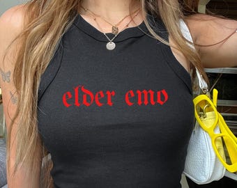 May include: A black sleeveless tank top featuring the text "elder emo" printed in a red gothic-style font across the chest. The garment has a high, rounded neckline and a fitted silhouette, showcasing a simple and clean design for casual wear.