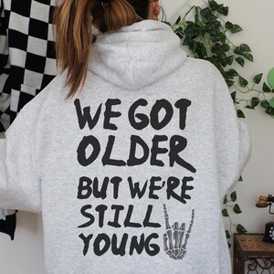 Peut inclure: Un sweat à capuche gris clair avec l'inscription noire "WE GOT OLDER BUT WE'RE STILL YOUNG" et un symbole de main de squelette. Le sweat à capuche est de style décontracté.