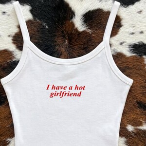 May include: A white sleeveless crop top with thin shoulder straps featuring the red embroidered text I have a hot girlfriend centered on the chest. The garment is made of a plain white fabric and includes the word Embroidered in the corner.