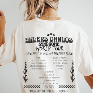 Puede incluir: Camiseta blanca con texto negro que dice "Ehlers Danlos Syndrome World Tour" y "Some days I'm a 10... on the pain scale". El texto enumera los síntomas del síndrome, incluyendo dolor, dislocaciones, subluxaciones, hipermovilidad, osteoartritis, esguinces, problemas gastrointestinales, cicatrices atróficas, y más. El texto también incluye "Est. since forever" y "Bendy Bitch Disorder".