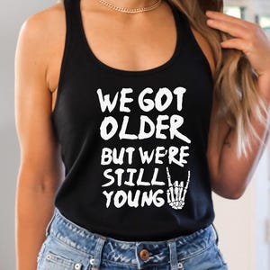 May include: A black racerback tank top featuring white text that reads WE GOT OLDER BUT WE'RE STILL YOUNG, accompanied by a graphic of a skeleton hand making a rock and roll sign. The tank top is paired with blue denim jeans.