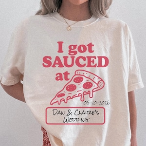 May include: Ivory-colored t-shirt with the text "I got SAUCED at" above a pizza slice graphic. Below the graphic is the date "05-10-2026" and the text "Dan & Claire's WEDDING" in a red rectangular box. The shirt is a casual style.