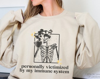 May include: A beige sweatshirt featuring a graphic of a skeleton holding a cup of coffee. The text "personally victimized by my immune system" is printed below the graphic.