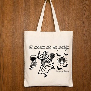 Op de afbeelding: Een crèmekleurige canvas draagtas met een zwarte grafische print. Het ontwerp bevat de tekst 'til death do us party' en 'Claire's Bach', een skelet met een heksenhoed en jurk, een wijnglas, een discobal en vleermuizen.