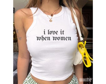 May include: A white sleeveless tank top featuring the black text i love it when women printed in a gothic font across the chest. The garment has a round neckline and a fitted silhouette, showcasing a clean design with no additional patterns or graphics.