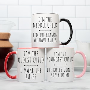 May include: Three white ceramic mugs with black text and red, pink, and black handles. The mugs have different sayings about being the oldest, middle, and youngest child. The text on the mugs reads: "I'm the oldest child, I make the rules", "I'm the middle child, I'm the reason we have rules", and "I'm the youngest child, the rules don't apply to me".