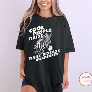 K&ouml;nnte beinhalten: Schwarzes T-Shirt mit wei&szlig;em Text, der "Cool People Raise Rare Disease Awareness" lautet. Neben dem Text ist ein Zebrakopf abgebildet.