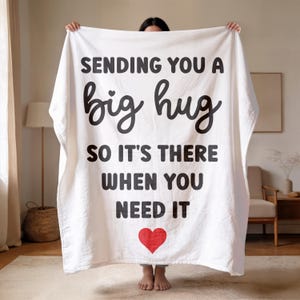 May include: White blanket with the text "SENDING YOU A big hug SO IT'S THERE WHEN YOU NEED IT" in black, with a red heart at the bottom. The blanket is held up by a person.