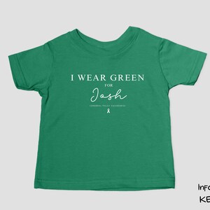 May include: A green infant t-shirt with the text "I Wear Green for Josh" in white. The shirt also includes the Cerebral Palsy Awareness ribbon. The shirt is a solid green colour.