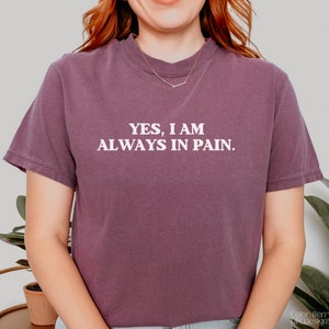 K&ouml;nnte beinhalten: Ein lilafarbenes, bauchfreies T-Shirt mit dem in Wei&szlig; gedruckten Text "YES, I AM ALWAYS IN PAIN."