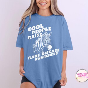 K&ouml;nnte beinhalten: Ein hellblaues T-Shirt mit wei&szlig;em Text, der "Cool People Raise Rare Disease Awareness" lautet. Hinter dem Text befindet sich eine wei&szlig;e Zebra-Illustration.