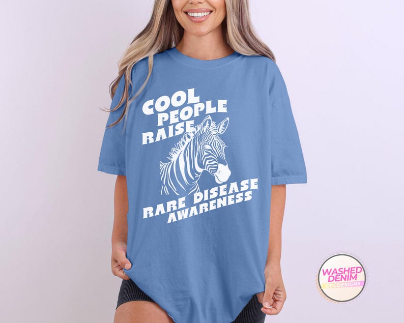K&ouml;nnte beinhalten: Ein hellblaues T-Shirt mit wei&szlig;em Text, der "Cool People Raise Rare Disease Awareness" lautet. Hinter dem Text befindet sich eine wei&szlig;e Zebra-Illustration.