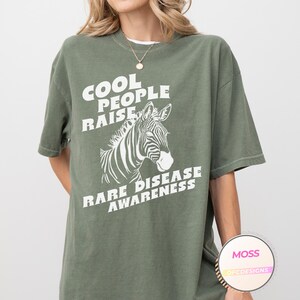 K&ouml;nnte beinhalten: Gr&uuml;nes T-Shirt mit wei&szlig;em Text, der "Cool People Raise Rare Disease Awareness" lautet, und einer Zebragrafik.