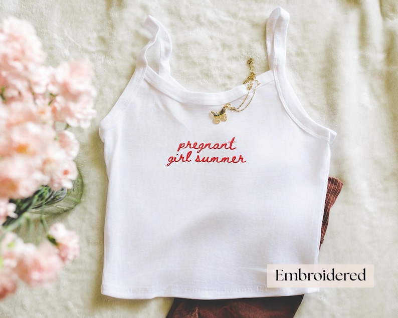 Puede incluir: Una camiseta de tirantes blanca con el texto bordado en rojo "pregnant girl summer" en el pecho. Un collar con colgante de mariposa en tono dorado descansa sobre el escote de la prenda. La palabra "Embroidered" aparece en un recuadro.