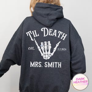 May include: A dark heather gray hooded sweatshirt featuring a white graphic on the back. The design displays a skeleton hand making a shaka sign with a ring, the text "TIL DEATH", "est. 2.2.2024", and the name "MRS. SMITH" in a distressed font.
