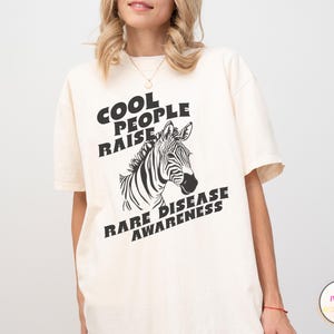 K&ouml;nnte beinhalten: Ein wei&szlig;es T-Shirt mit schwarzem Text, der "Cool People Raise Rare Disease Awareness" lautet, und einer Illustration eines schwarz-wei&szlig;en Zebras.