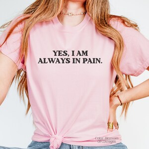 K&ouml;nnte beinhalten: Ein pinkfarbenes T-Shirt mit dem Text "YES, I AM ALWAYS IN PAIN." in Schwarz gedruckt.