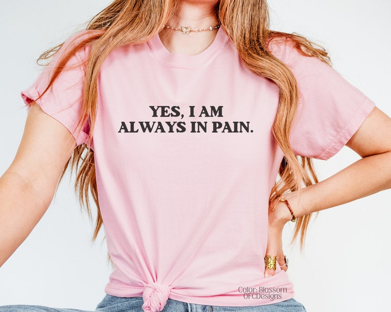 K&ouml;nnte beinhalten: Ein pinkfarbenes T-Shirt mit dem Text "YES, I AM ALWAYS IN PAIN." in Schwarz gedruckt.