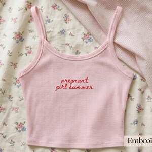 Puede incluir: Una camiseta de tirantes acanalada de color rosa claro con tirantes finos y escote redondo. El centro de la prenda muestra el texto bordado en rojo "pregnant girl summer" en una fuente cursiva, ofreciendo un dise&ntilde;o claro y legible.