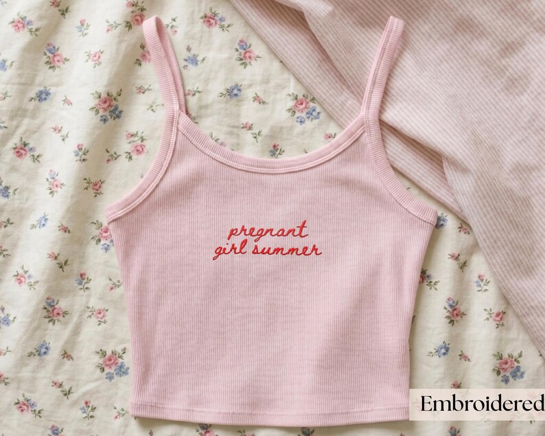 Puede incluir: Una camiseta de tirantes acanalada de color rosa claro con tirantes finos y escote redondo. El centro de la prenda muestra el texto bordado en rojo "pregnant girl summer" en una fuente cursiva, ofreciendo un dise&ntilde;o claro y legible.