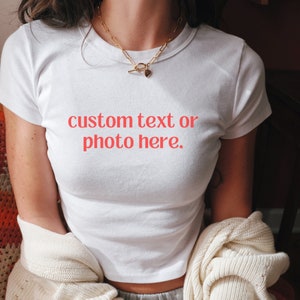 May include: A white crop top with the text "custom text or photo here" in red. The shirt is short-sleeved and has a crew neck. A gold-colored necklace with a heart pendant is worn. A cream-colored sweater is draped over the arms.