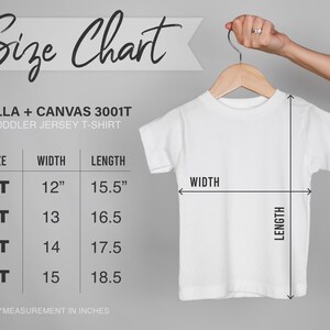 May include: White toddler jersey t-shirt hanging on a wooden hanger. The image includes a size chart with measurements in inches for sizes 2T to 5T. The chart shows the width and length of the shirts. The text on the chart reads "Size Chart" and "BELLA + CANVAS 3001T TODDLER JERSEY T-SHIRT."