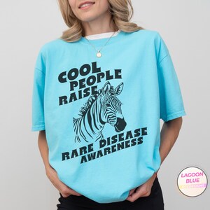 K&ouml;nnte beinhalten: Ein hellblaues T-Shirt mit einem schwarzen Zebra-Motiv und dem Text "Cool People Raise Rare Disease Awareness".